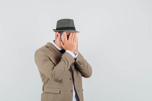 Hombre joven mirando a través de los dedos en traje, sombrero y mirando asustado, vista frontal.