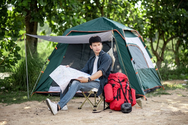 Hombre joven mochilero sentado frente a la carpa en el bosque natural y mirando en el mapa de papel de los senderos del bosque para planificar mientras acampa el viaje en las vacaciones de verano, espacio de copia