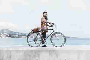 Foto gratuita hombre joven de moda con su bicicleta de pie en un rompeolas cerca de la costa