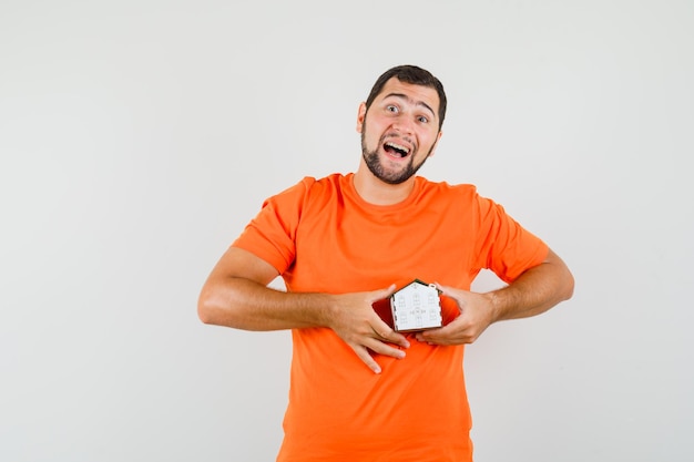 Hombre joven con modelo de casa en camiseta naranja y mirando feliz, vista frontal.
