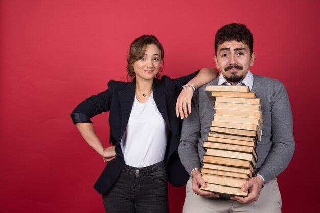 Hombre joven con montón de libros y colega de pie en la pared roja