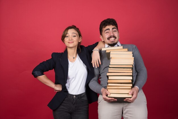 Hombre joven con montón de libros y colega de pie en la pared roja