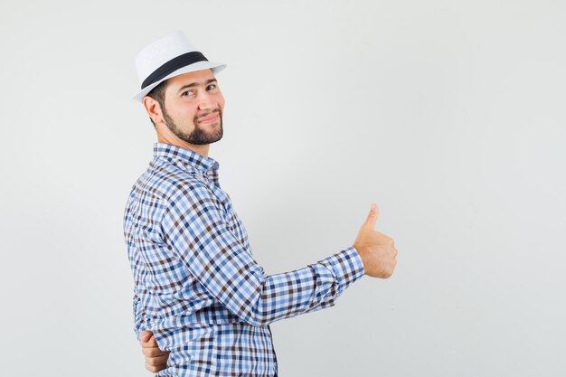 Hombre joven mostrando el pulgar hacia arriba en camisa a cuadros, sombrero y mirando alegre. .