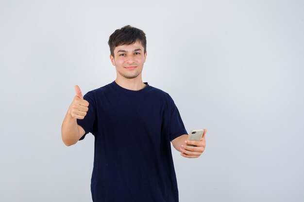 Hombre joven mostrando el pulgar hacia arriba, sosteniendo el teléfono móvil en camiseta negra y mirando confiado. vista frontal.