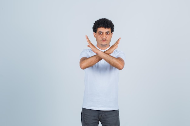 Hombre joven mostrando restricción o gesto x en camiseta blanca y jeans y mirando serio. vista frontal.