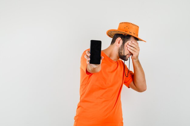 Hombre joven mostrando teléfono móvil en camiseta naranja, sombrero y mirando asustado. vista frontal.