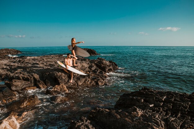 Hombre joven y mujer apuntando al lado con tablas de surf en roca cerca del mar