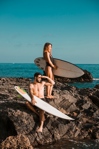 Hombre joven y mujer sorprendida con tablas de surf en roca cerca del mar