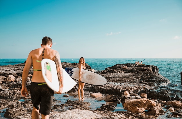 Hombre joven y mujer con tablas de surf en la orilla de piedra cerca del agua