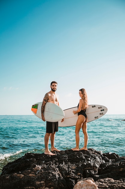 Hombre joven y mujer con tablas de surf en piedra cerca del agua