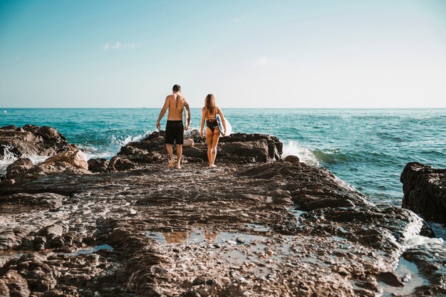 Hombre joven y mujer con tablas de surf que van en la orilla de piedra al agua