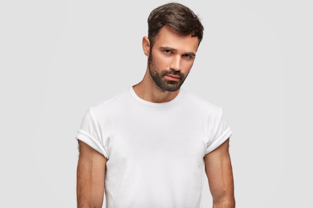 Hombre joven musculoso serio con barba oscura, cabello, vestido con camiseta blanca casual, tiene cuerpo musculoso, escucha atentamente algo, aislado sobre una pared blanca. Chico sin afeitar se encuentra en el interior