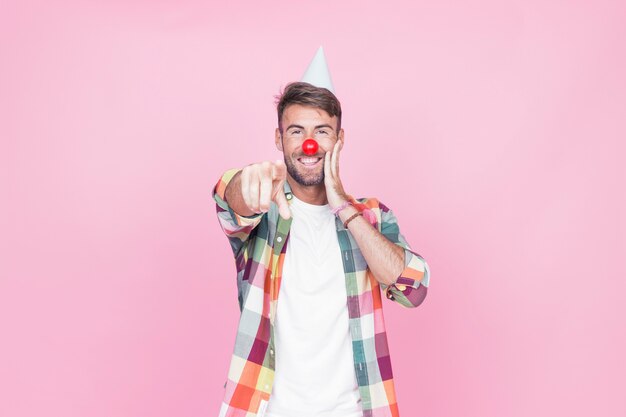 Hombre joven con nariz de payaso apuntando su dedo sobre fondo rosa