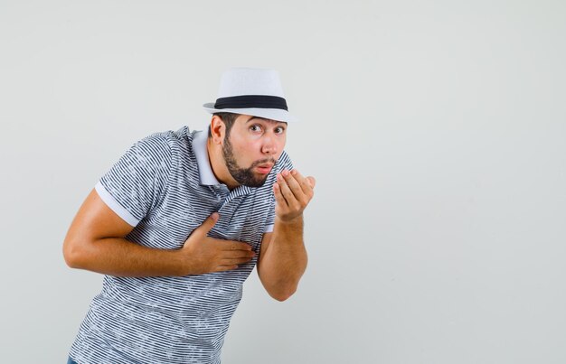 Hombre joven con náuseas en camiseta, sombrero y aspecto enfermo, vista frontal.