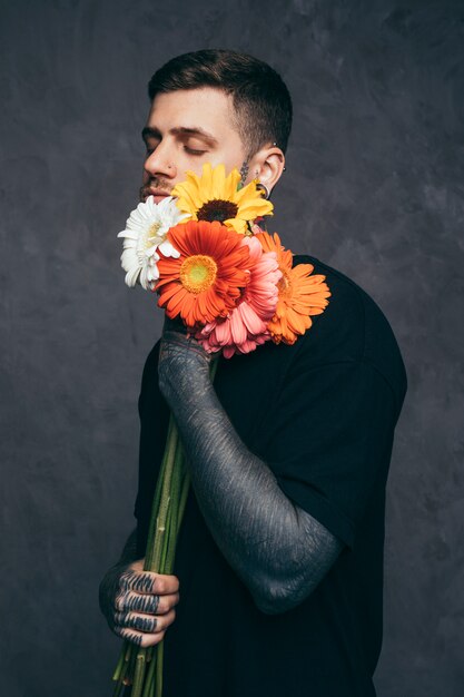 Hombre joven con los ojos cerrados y tatuado en su mano sosteniendo una flor de gerbera en la mano
