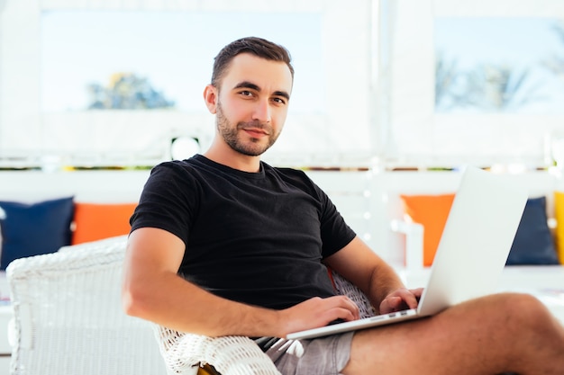 Hombre joven con ordenador portátil en la cafetería al aire libre en las vacaciones de verano