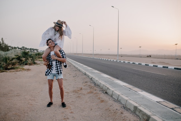 Hombre joven en pantalones cortos de mezclilla con su elegante novia sobre los hombros de pie cerca de la carretera. Adorable mujer en blusa blanca vintage pasar tiempo con su novio y divertirse en una cita al aire libre