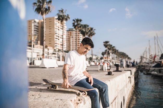 Foto gratuita hombre joven con una patineta sentado en el muro de contención