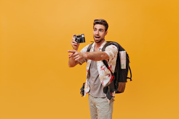 Hombre joven con peinado fresco y barba en elegante camisa de verano posando con cámara y mochila y mostrando a un lado su dedo