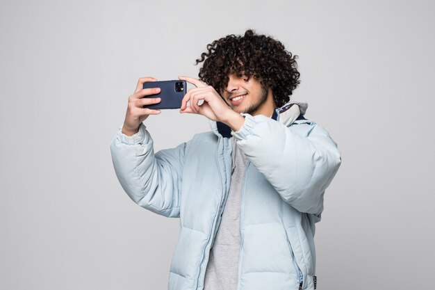 Hombre joven con pelo rizado mediante teléfono móvil sobre pared blanca aislada