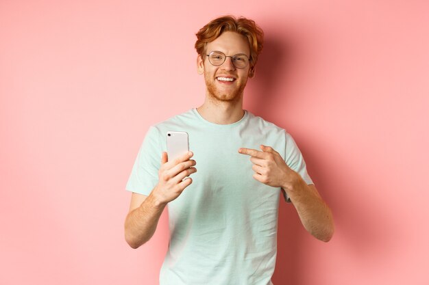Hombre joven con pelo rojo y barba, con camiseta y gafas, sonriendo mientras señala con el dedo al teléfono inteligente, recomienda promoción en línea, fondo rosa.