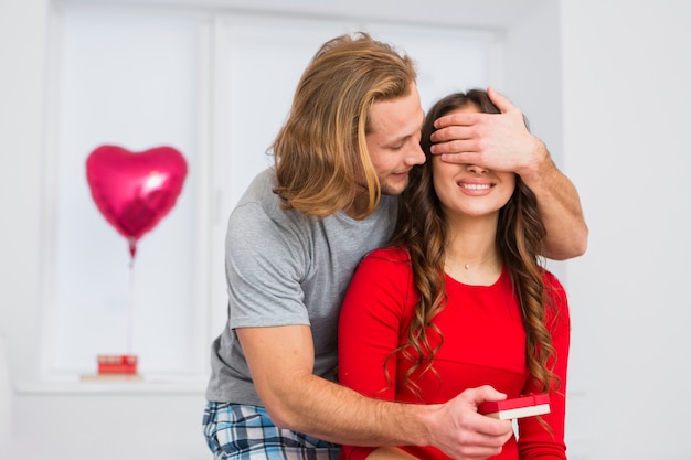 Hombre joven del pelo rubio que cubre los ojos de su novia mientras que da el regalo a ella