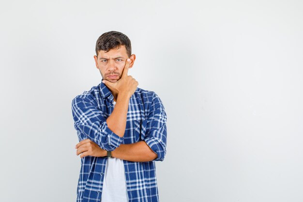 Hombre joven pensando con los dedos en la mejilla y la barbilla en camisa y mirando triste, vista frontal.