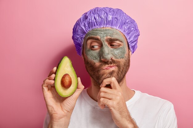 Hombre joven pensativo con máscara de arcilla en la cara, sostiene una rodaja de aguacate, recibe tratamientos de spa, sostiene la barbilla, tiene rastrojo, usa gorro de baño, camiseta blanca