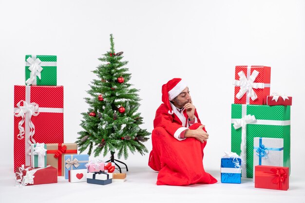 Hombre joven pensativo vestido como Papá Noel con regalos y árbol de Navidad decorado sentado en el suelo sobre fondo blanco.