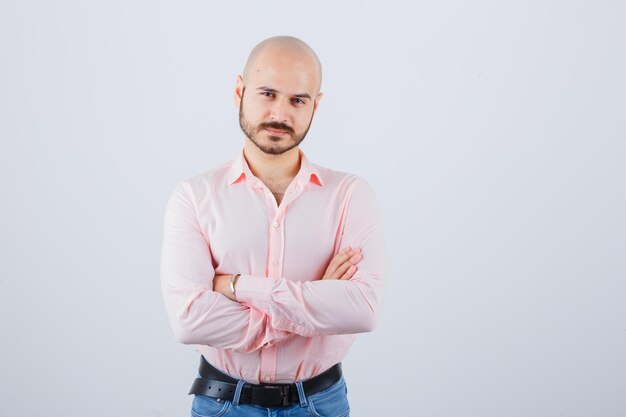 Hombre joven de pie con los brazos cruzados en camisa rosa, jeans y mirando confiado. vista frontal.