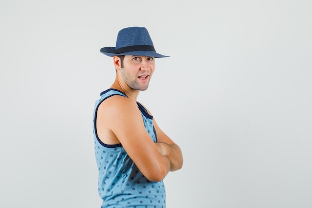 Hombre joven de pie con los brazos cruzados en camiseta azul, sombrero y mirando confiado.