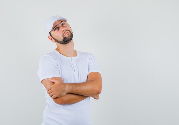 Hombre joven de pie con los brazos cruzados en camiseta blanca, gorra y mirando pensativo, vista frontal. espacio para texto