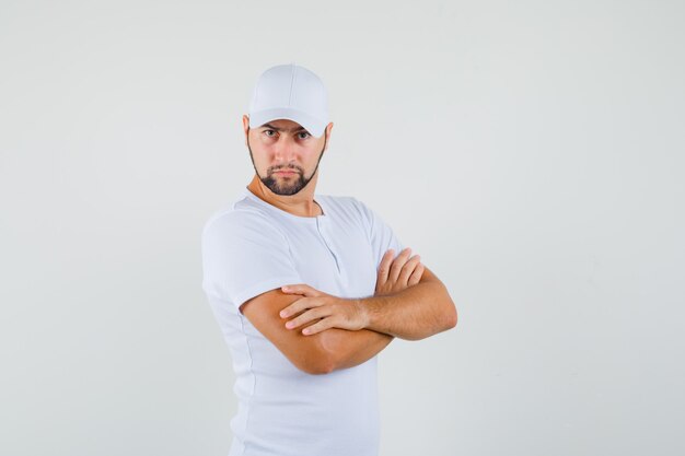 Hombre joven de pie con los brazos cruzados en camiseta blanca y mirando estricto. vista frontal.