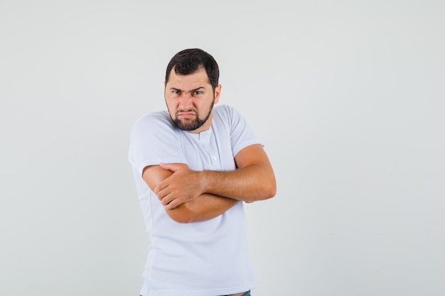 Hombre joven de pie con los brazos cruzados en camiseta blanca y mirando ofendido, vista frontal.