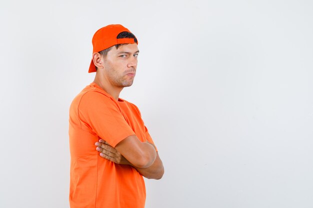 Hombre joven de pie con los brazos cruzados en camiseta naranja y gorra y mirando astuto