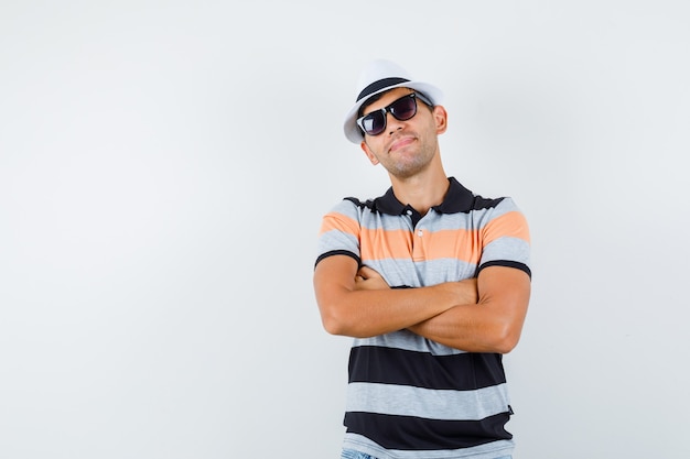 Hombre joven de pie con los brazos cruzados en camiseta y sombrero y luciendo bien