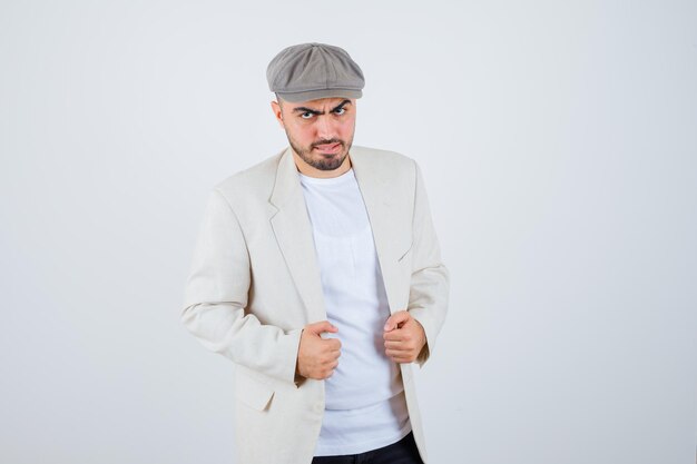 Hombre joven de pie con la espalda recta y posando al frente con camiseta blanca, chaqueta y gorra gris y mirando serio