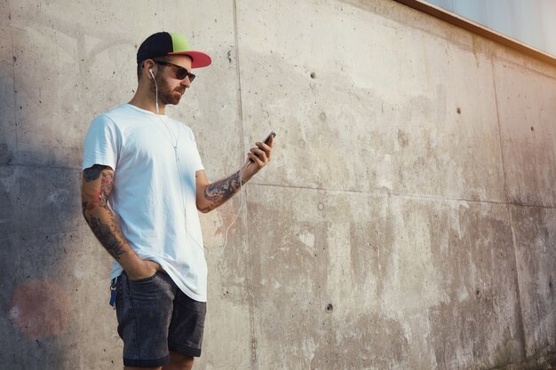 Hombre joven de pie junto a una pared de hormigón gris mirando la pantalla de su teléfono inteligente y escuchando música en sus tapones para los oídos blancos