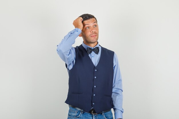 Hombre joven de pie con la mano en la cabeza en traje, jeans y mirando pensativo.