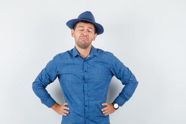 Hombre joven de pie con las manos en la cintura en camisa azul, sombrero y con cara de tristeza. vista frontal.