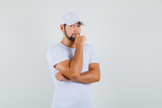 Hombre joven de pie mientras apoya la barbilla en la mano en camiseta, gorra y parece preocupado. vista frontal.