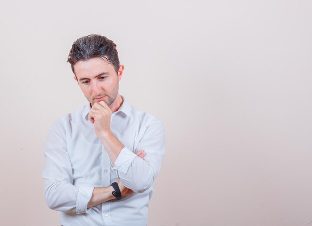 Hombre joven de pie en pose de pensamiento en camisa blanca y mirando inteligente