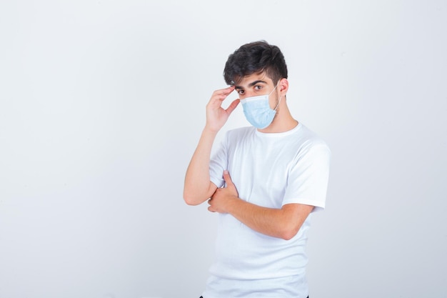 Hombre joven de pie en pose de pensamiento en camiseta blanca, máscara y aspecto sensible