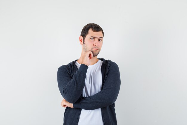 Hombre joven de pie en pose de pensamiento en camiseta blanca y sudadera con capucha negra con cremallera frontal y mirando serio, vista frontal.