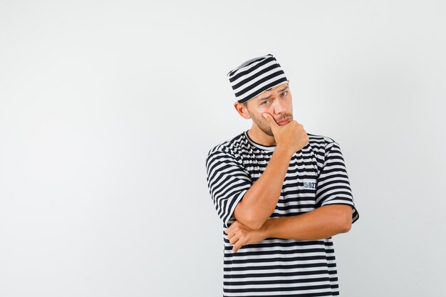 Hombre joven de pie en pose de pensamiento en camiseta a rayas, sombrero y aspecto sensato.