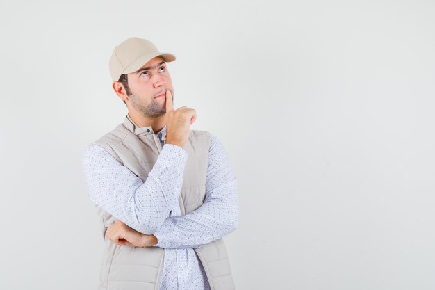 Hombre joven de pie en pose de pensamiento, mirando a otro lado y poniendo el dedo índice en la boca con chaqueta beige y gorra y mirando pensativo, vista frontal.