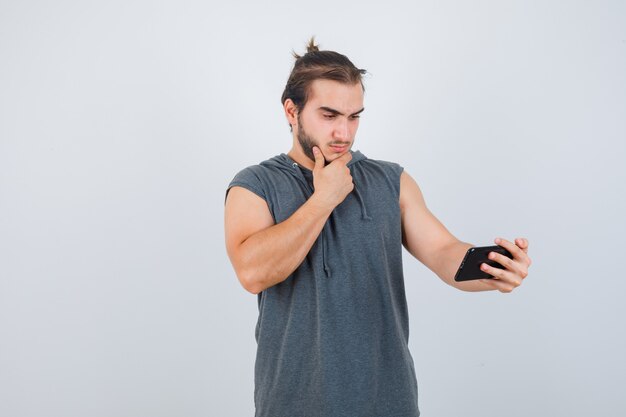 Hombre joven de pie en pose de pensamiento, sosteniendo el teléfono en una camiseta con capucha y mirando sensible, vista frontal.