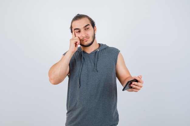 Hombre joven de pie en pose de pensamiento, sosteniendo el teléfono en la mano con una camiseta con capucha y luciendo sensato, vista frontal.