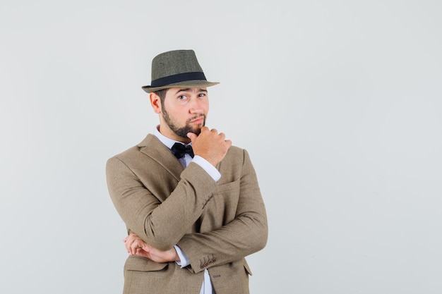 Hombre joven de pie en pose de pensamiento en traje, sombrero y aspecto sensato. vista frontal.