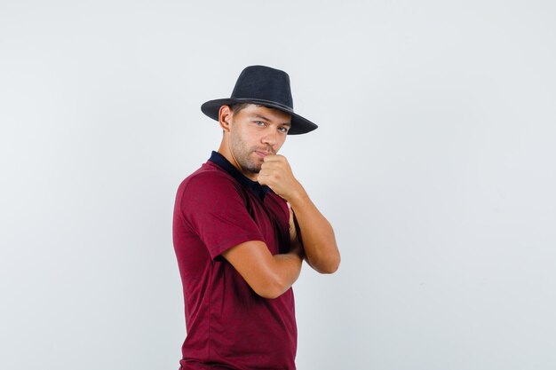Hombre joven de pie con el puño levantado en camiseta, sombrero y mirando confiado, vista frontal.